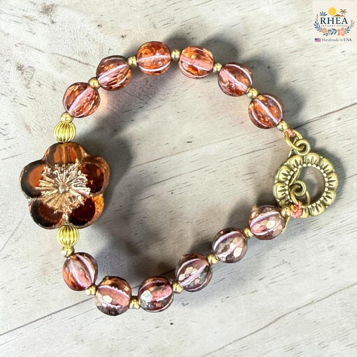 Rhea Artistry Studio handmade garden glass bead bracelet with floral focal element, brass hardware, and toggle clasp. Features warm amber and pink tones.