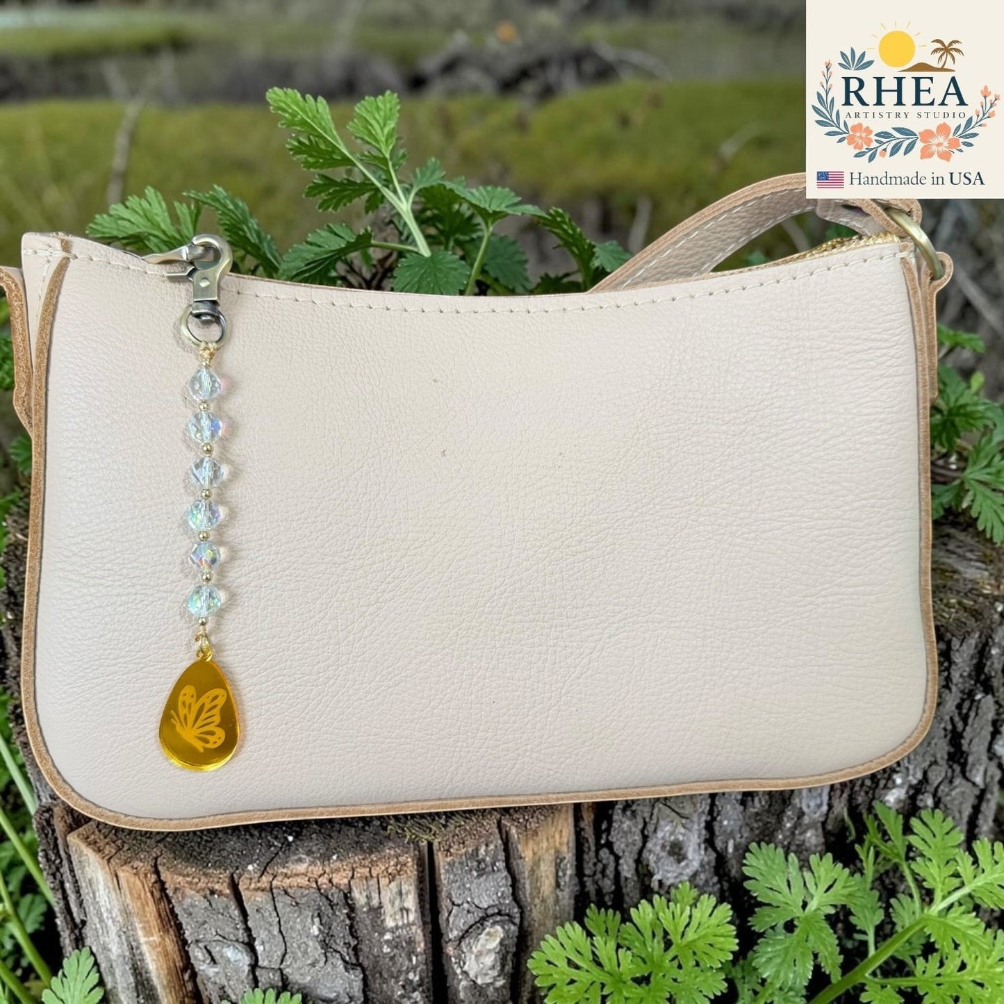 Rhea Artistry Studio cream leather bag with a handcrafted charm featuring multi-faceted glass beads and a gold butterfly pendant.