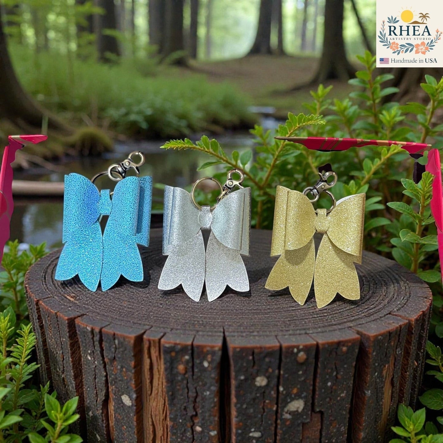 Rhea Artistry Studio handmade glitter bow bag charms in blue, silver, and gold with brass hardware, displayed on a wooden stump.