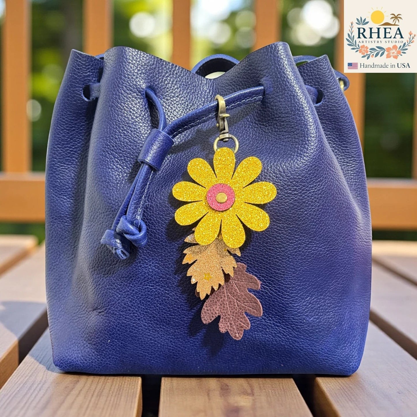 Rhea Artistry Studio yellow glitter flower charm with two leather leaves on a blue pebbled leather drawstring bag.