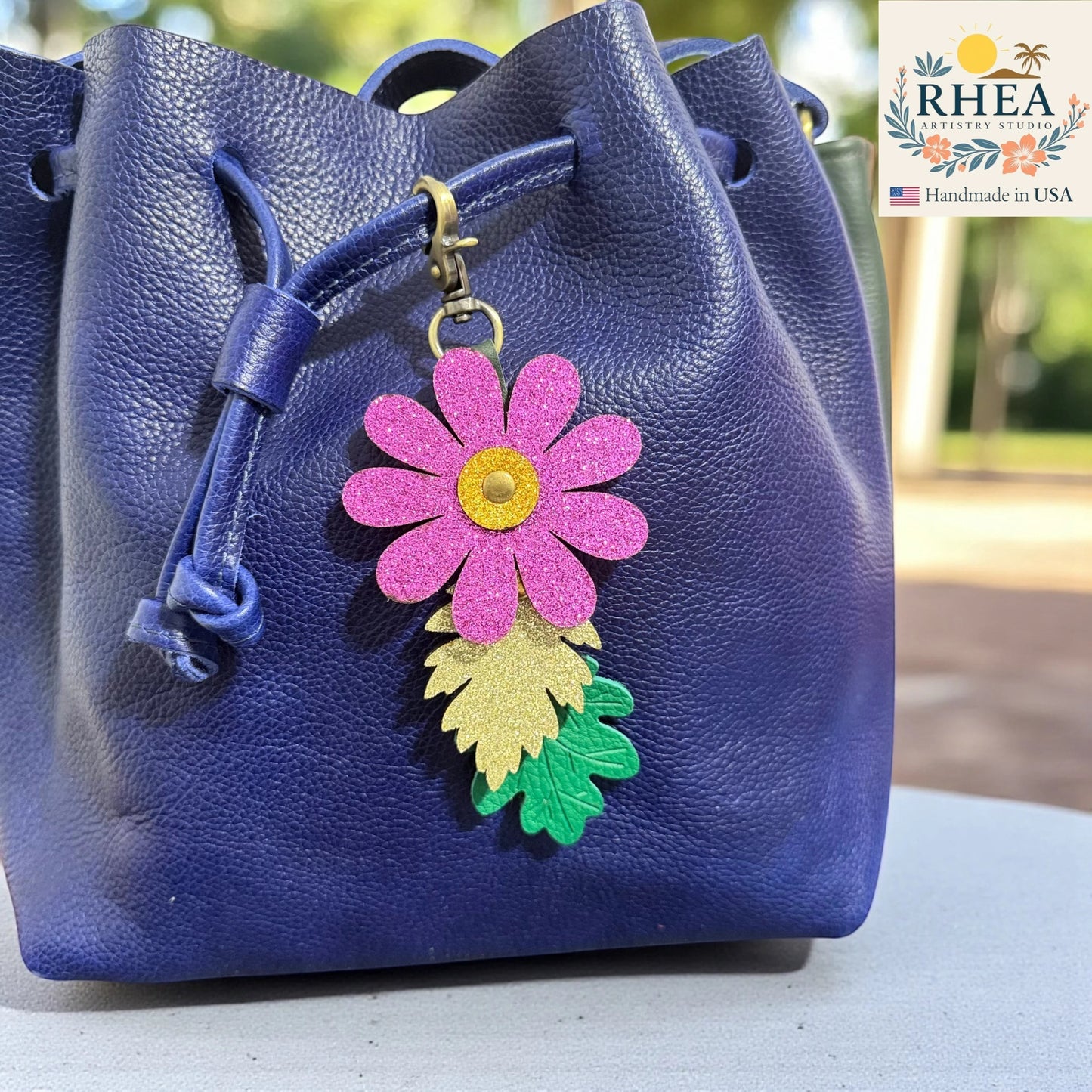 Rhea Artistry Studio pink glitter flower charm with gold center and two leather leaves, one glitter gold, one green embossed. Attached to a blue leather bucket bag.