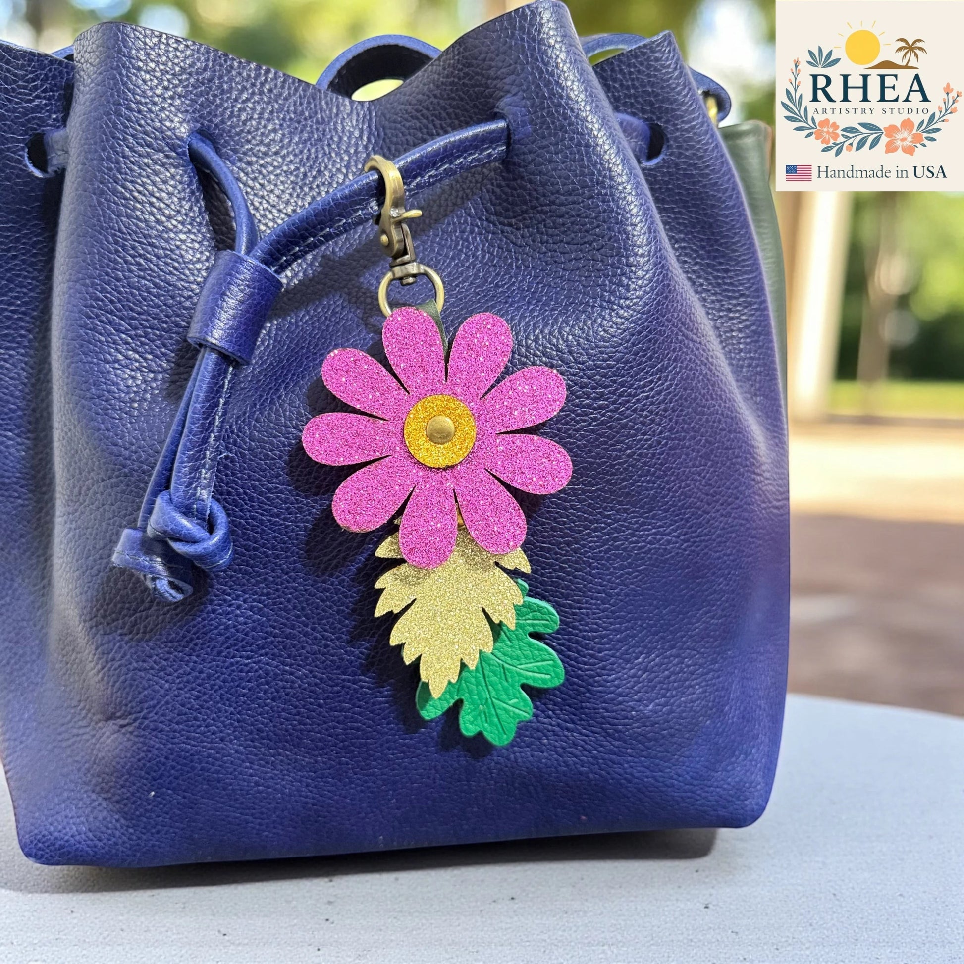 Rhea Artistry Studio pink glitter flower charm with gold center and two leather leaves, one glitter gold, one green embossed. Attached to a blue leather bucket bag.