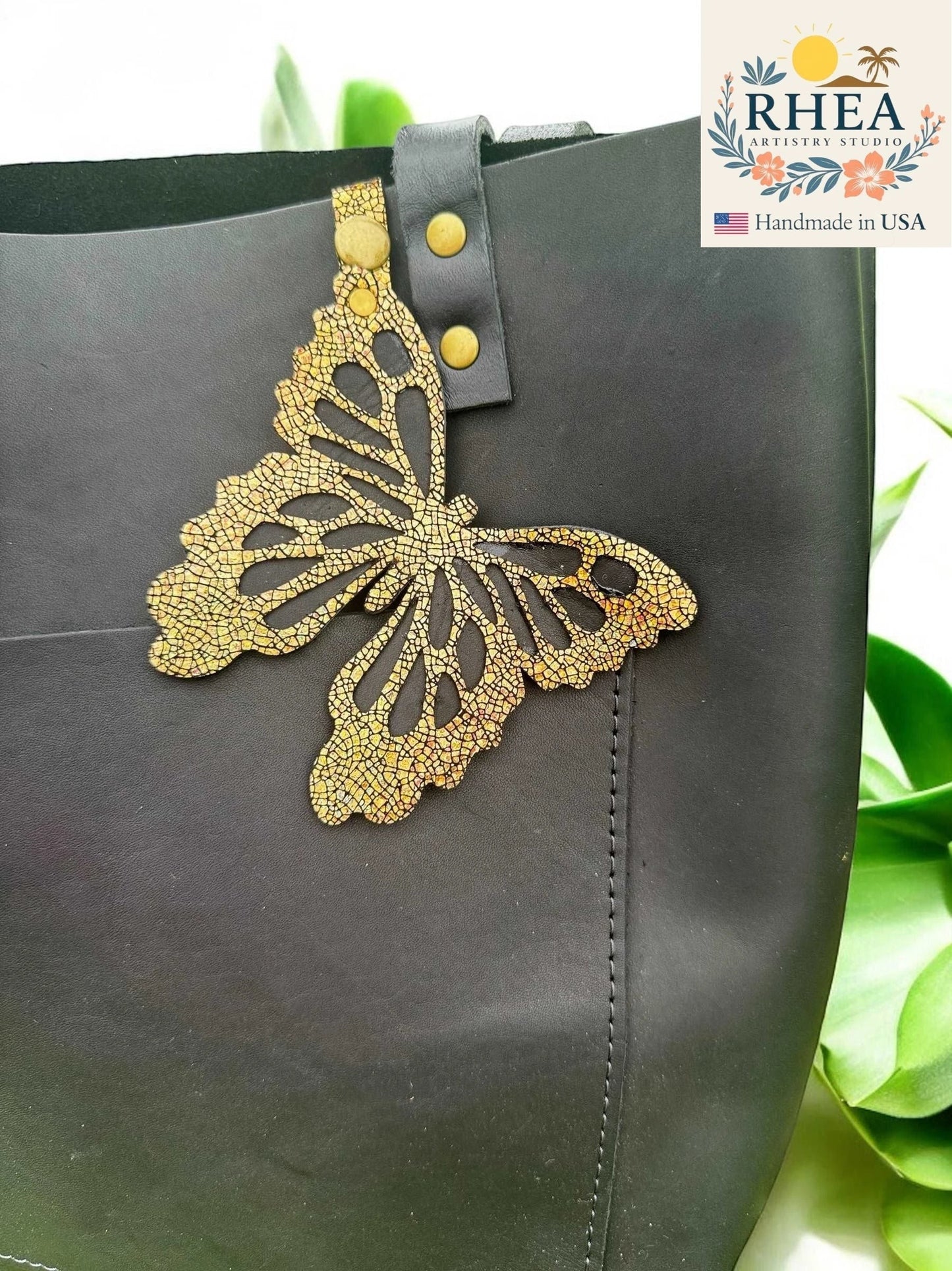 Rhea Artistry Studio gold metallic leather butterfly charm with iridescent finish, attached to a black tote bag.