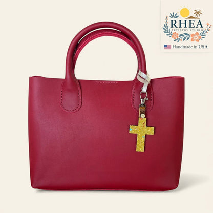 Rhea Artistry Studio red leather tote bag with a glitter gold cross charm attached to the handle.