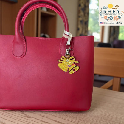 Rhea Artistry Studio handmade gold glitter bell charm with colorful accents, attached to a red leather handbag.