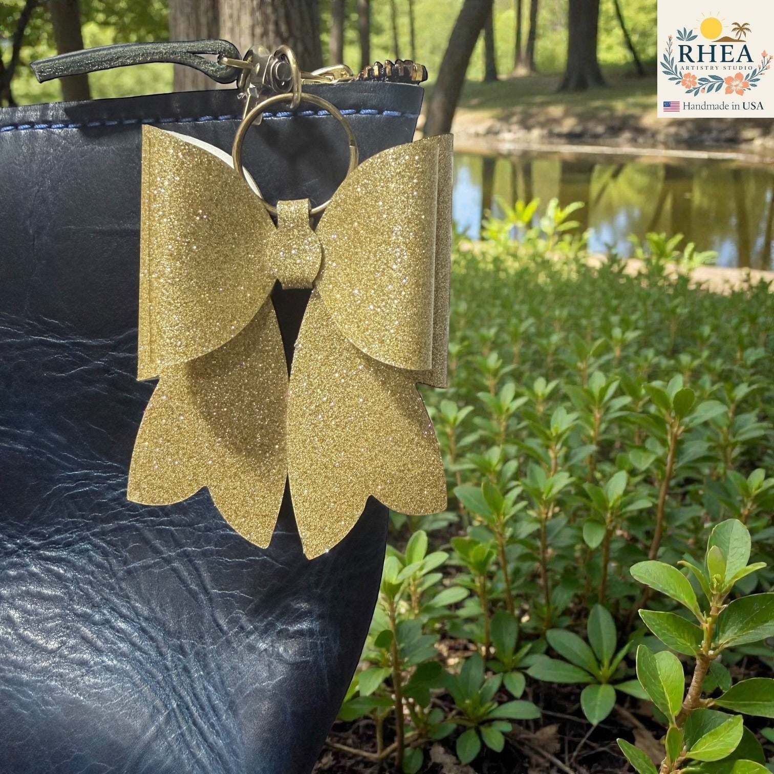 Rhea Artistry Studio gold glitter double layer French bow bag charm with brass hardware attached to a navy blue bag.