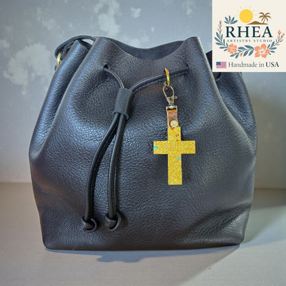 Rhea Artistry Studio gold glitter cross charm with colorful flecks, attached to a black leather drawstring bag.