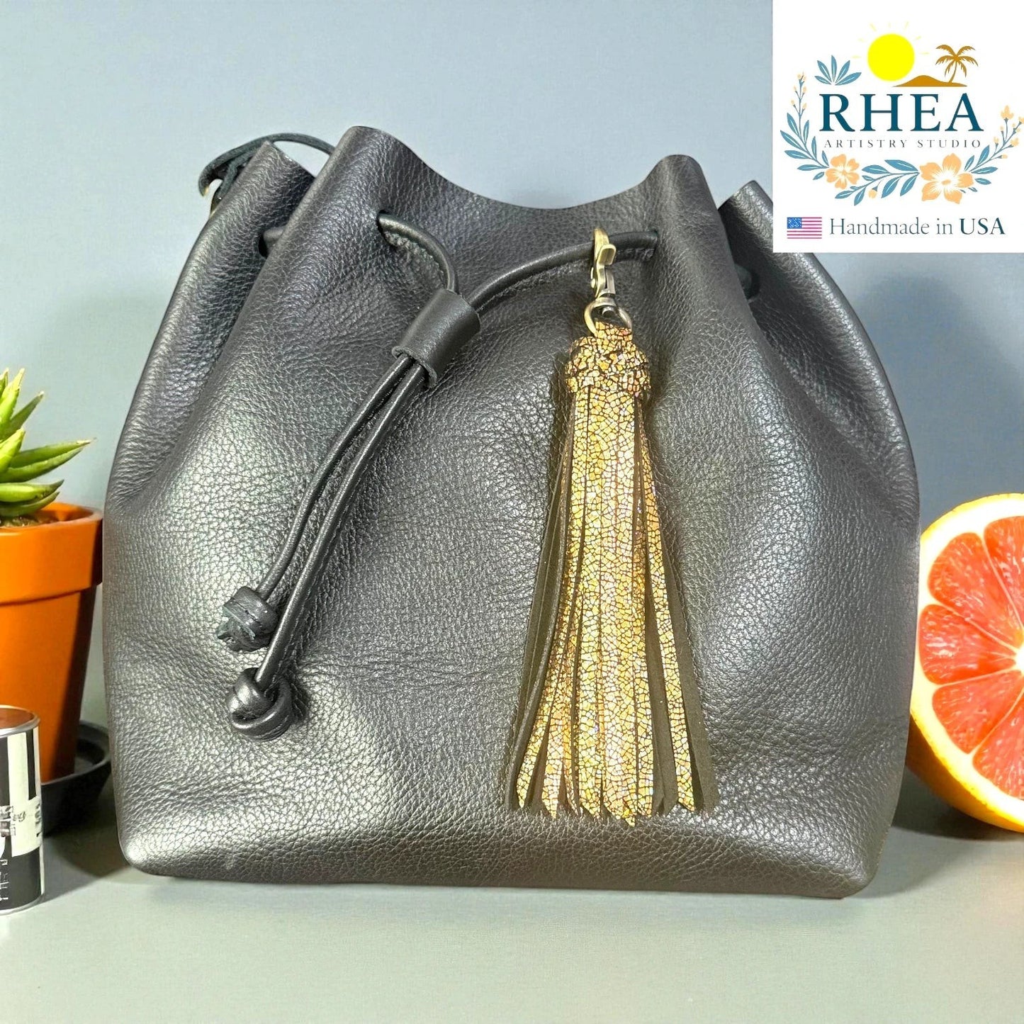 Rhea Artistry Studio black leather bucket bag with a gold glitter leather tassel charm and antique brass lobster clasp.