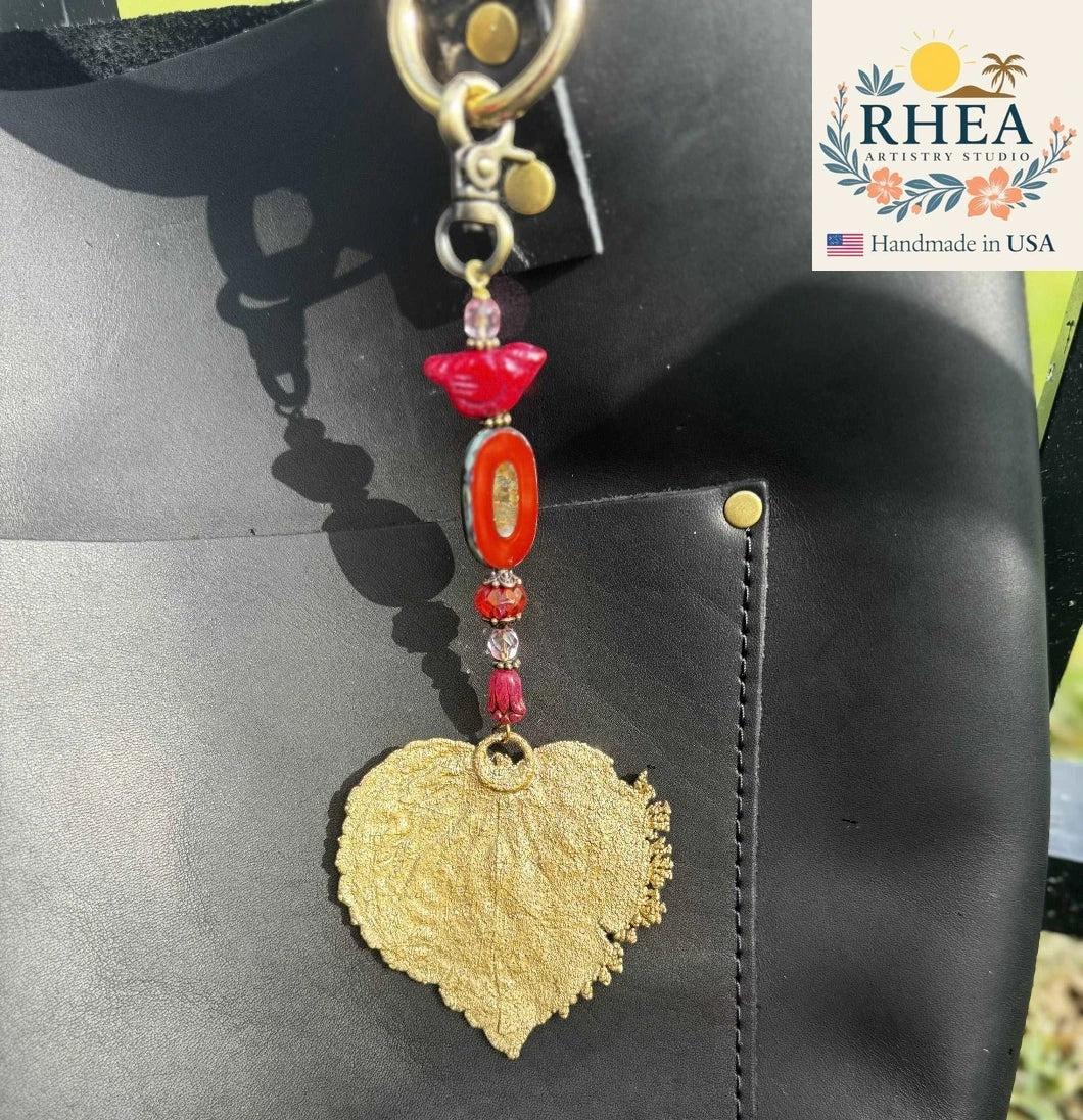 Rhea Artistry Studio gold leaf charm with red bird and bead accents, attached to a black leather bag.