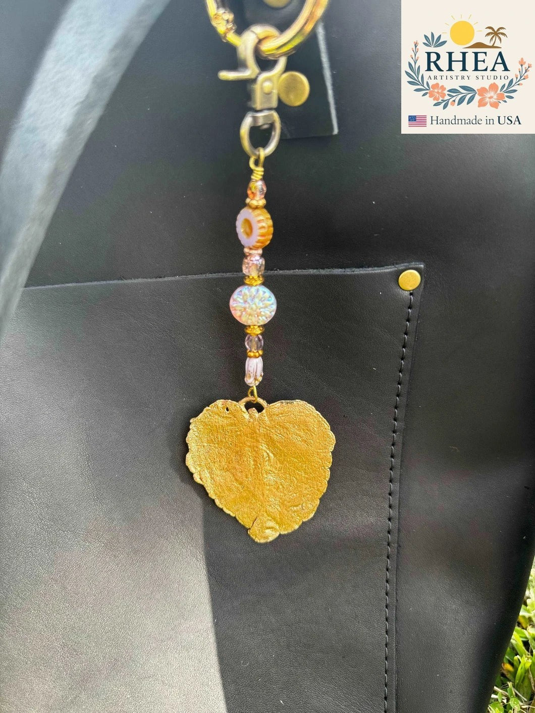 Rhea Artistry Studio gold-dipped hibiscus leaf charm with violet and iridescent beads on a black leather bag.