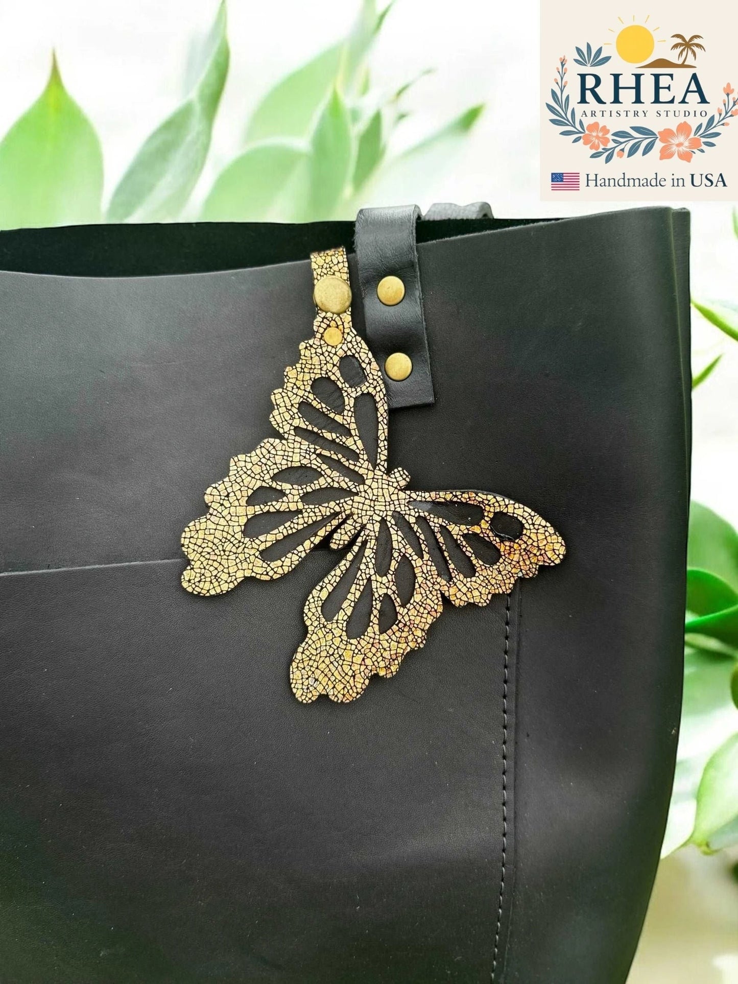 Rhea Artistry Studio gold cracked leather butterfly charm with metallic iridescent finish attached to a black tote bag.