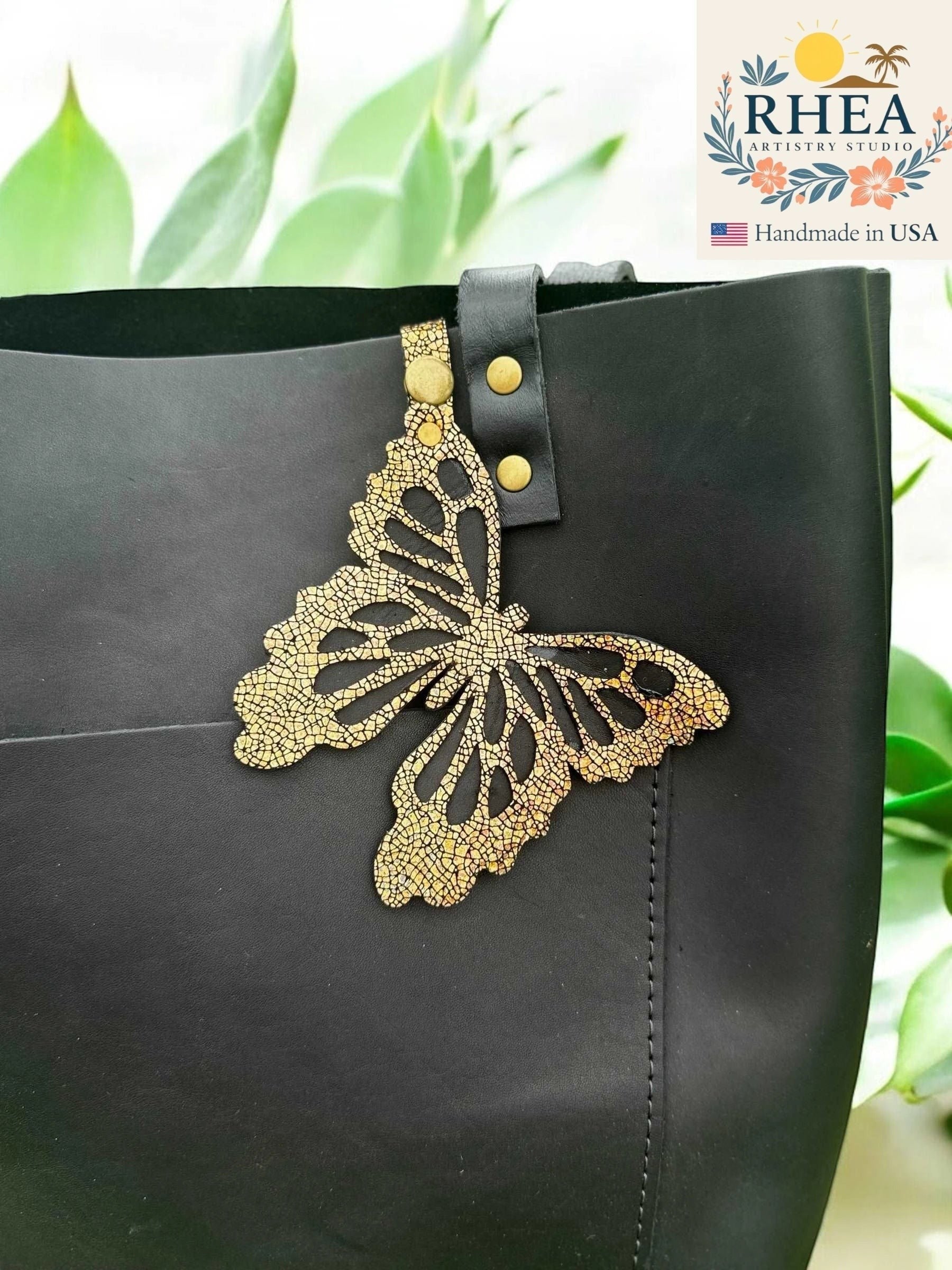 Rhea Artistry Studio gold cracked leather butterfly charm with metallic iridescent finish attached to a black tote bag.