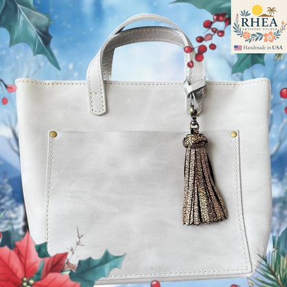 Rhea Artistry Studio handmade chunky leather tassel bag charm in gold metallic finish with lobster clasp attached to a light grey tote bag.