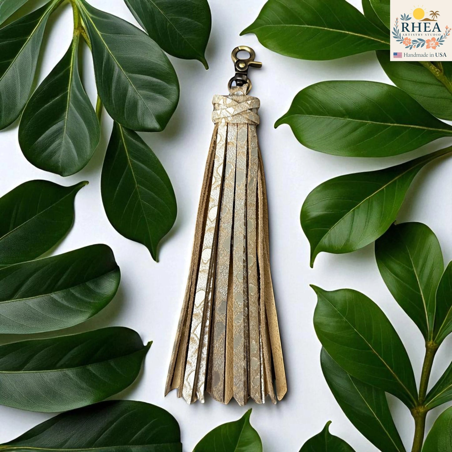 Rhea Artistry Studio handmade large gold leather tassel keychain with lobster clasp, surrounded by green leaves.