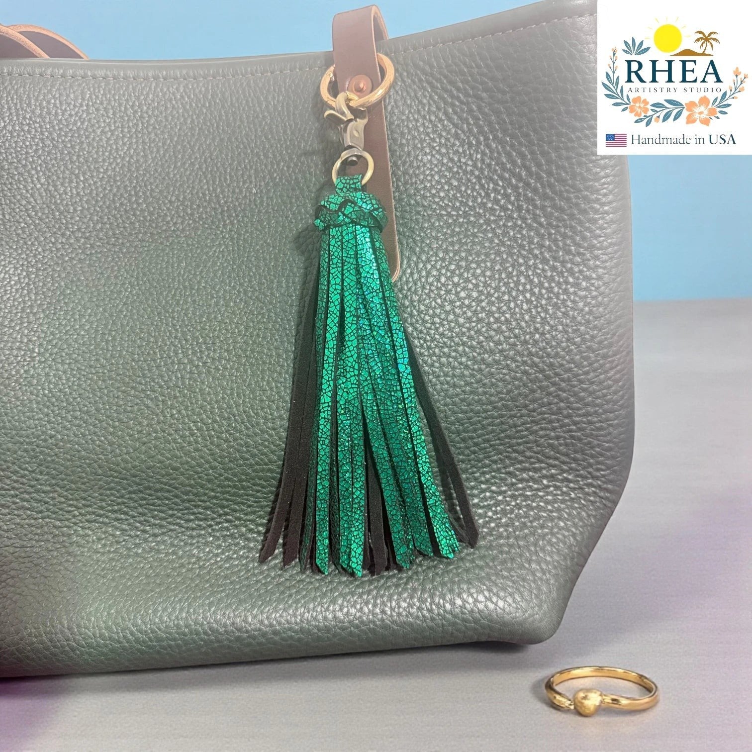 Rhea Artistry Studio handmade green leather tassel keychain with lobster clasp attached to a grey pebbled leather tote bag.
