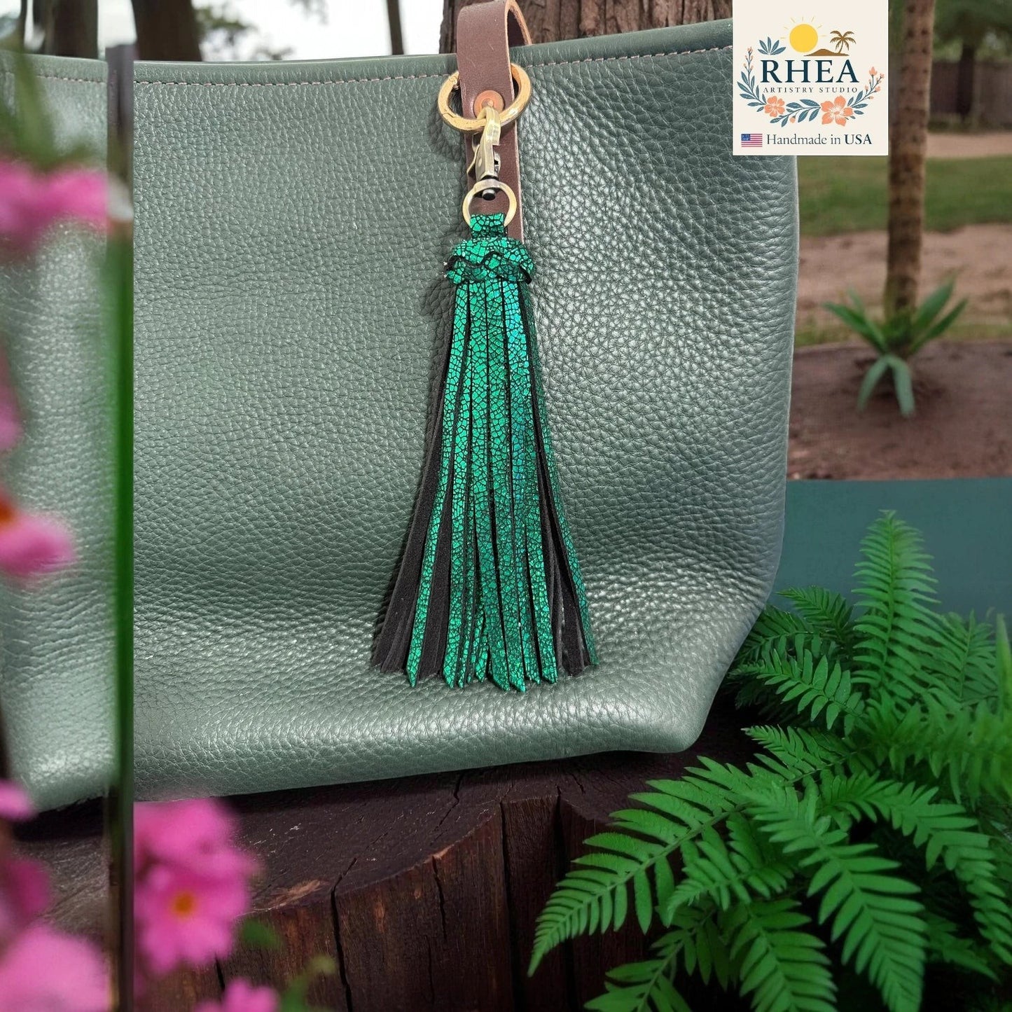 Rhea Artistry Studio dual-tone green leather tassel keychain with glitter finish and antique brass clasp attached to a green pebbled leather tote bag.