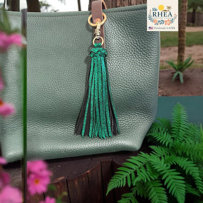 Rhea Artistry Studio dual-tone green leather tassel keychain with glitter finish and antique brass clasp attached to a green pebbled leather tote bag.