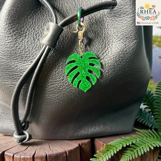 Rhea Artistry Studio green glitter monstera leaf acrylic charm with antique brass clasp on a black leather bag.