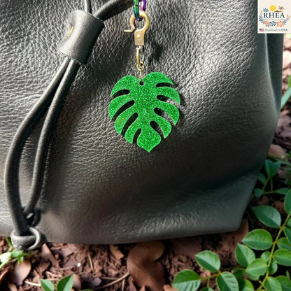 Rhea Artistry Studio green glitter monstera leaf acrylic charm with antique brass clasp on black leather bag.