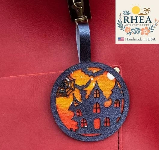 Rhea Artistry Studio handpainted Halloween charm featuring a haunted house silhouette with bats and spiderwebs against a sunset sky, made from leather.