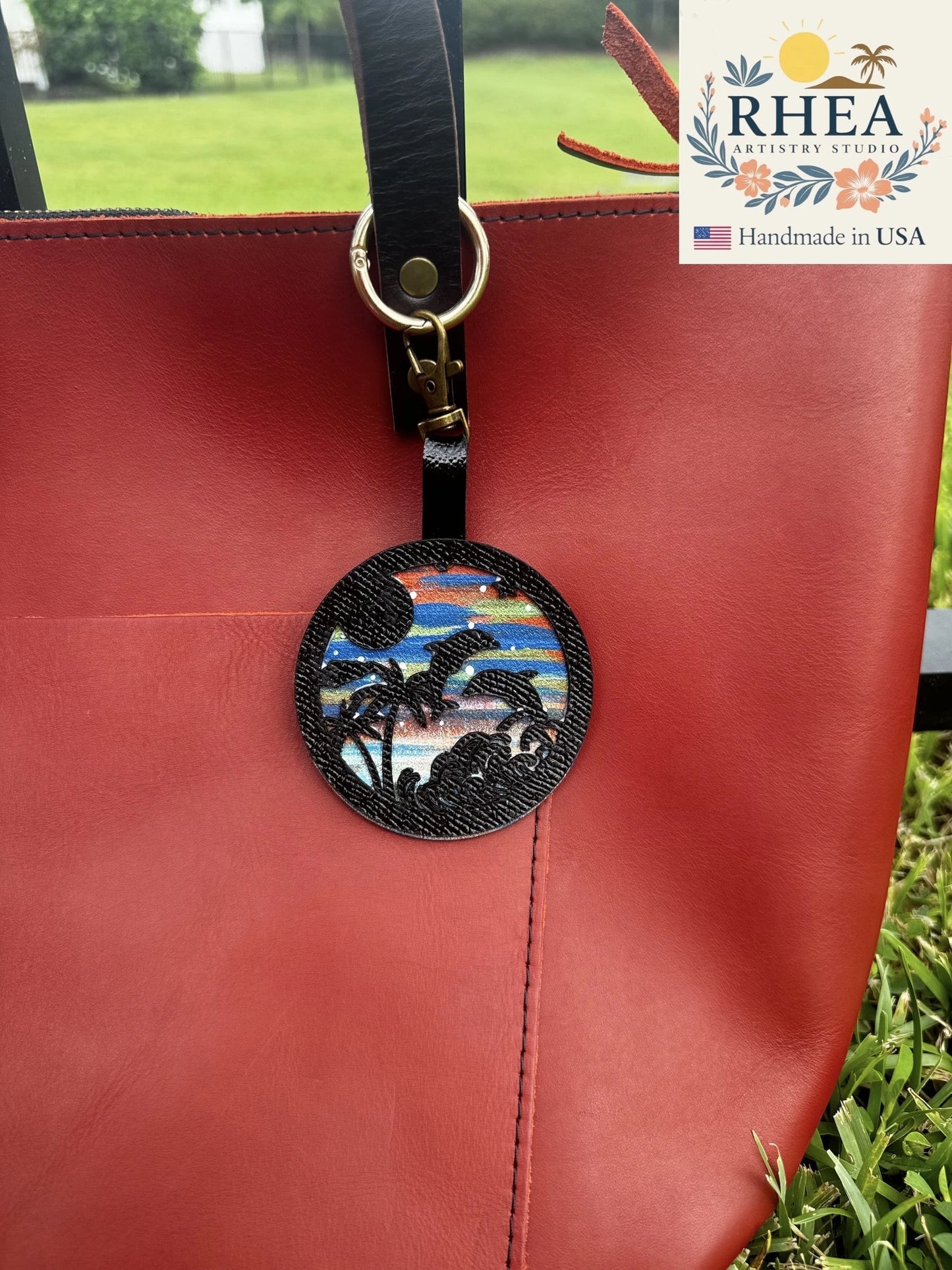 Rhea Artistry Studio hand-painted leather bag charm featuring dolphins, palm trees, and a colorful sunset scene.