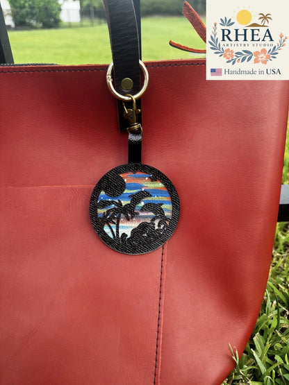 Rhea Artistry Studio hand-painted leather bag charm featuring dolphins, palm trees, and a colorful sunset scene.