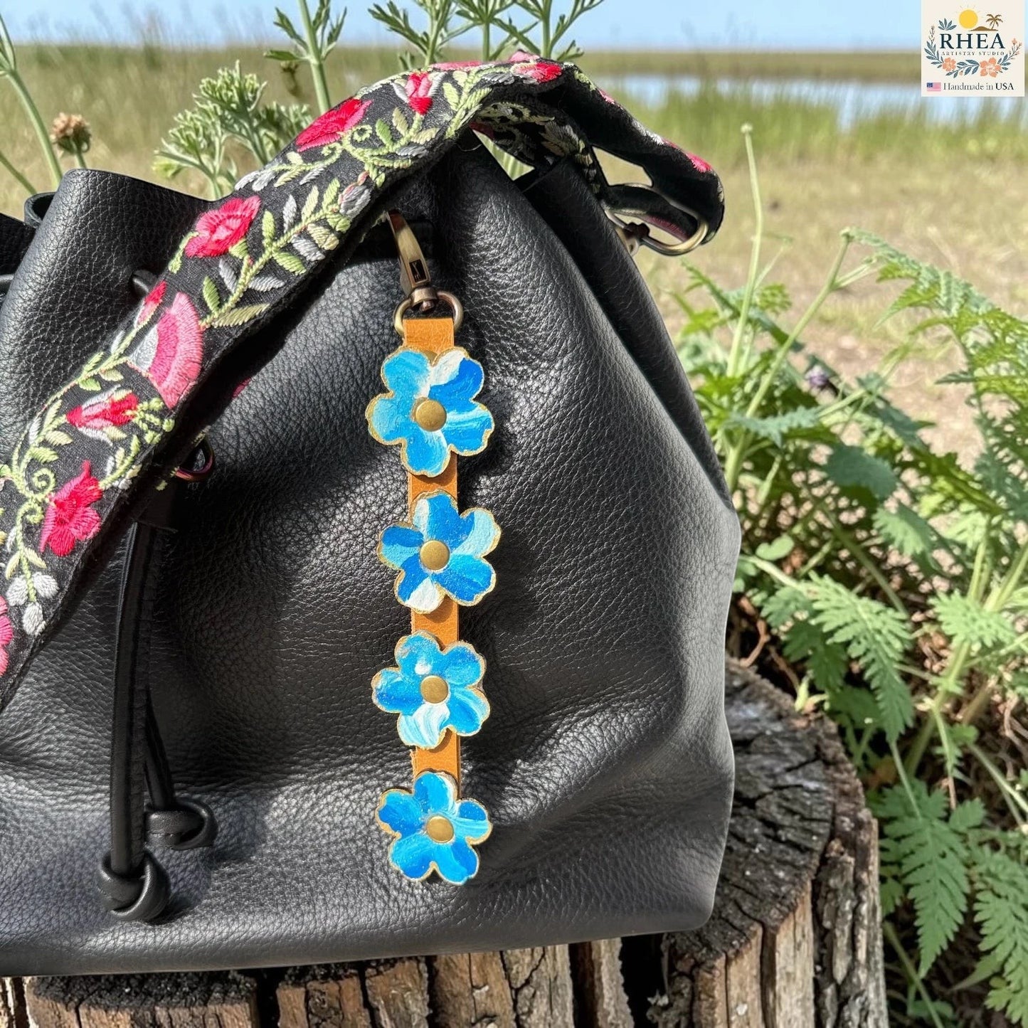 Rhea Artistry Studio Hand-Painted Leather Flower Bag Charm featuring organic painted leather flowers, brass rivets, honey leather strap, and antique brass clasp on a black bag