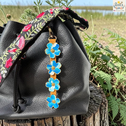 Rhea Artistry Studio Hand-Painted Leather Flower Bag Charm featuring organic painted leather flowers, brass rivets, honey leather strap, and antique brass clasp on a black bag