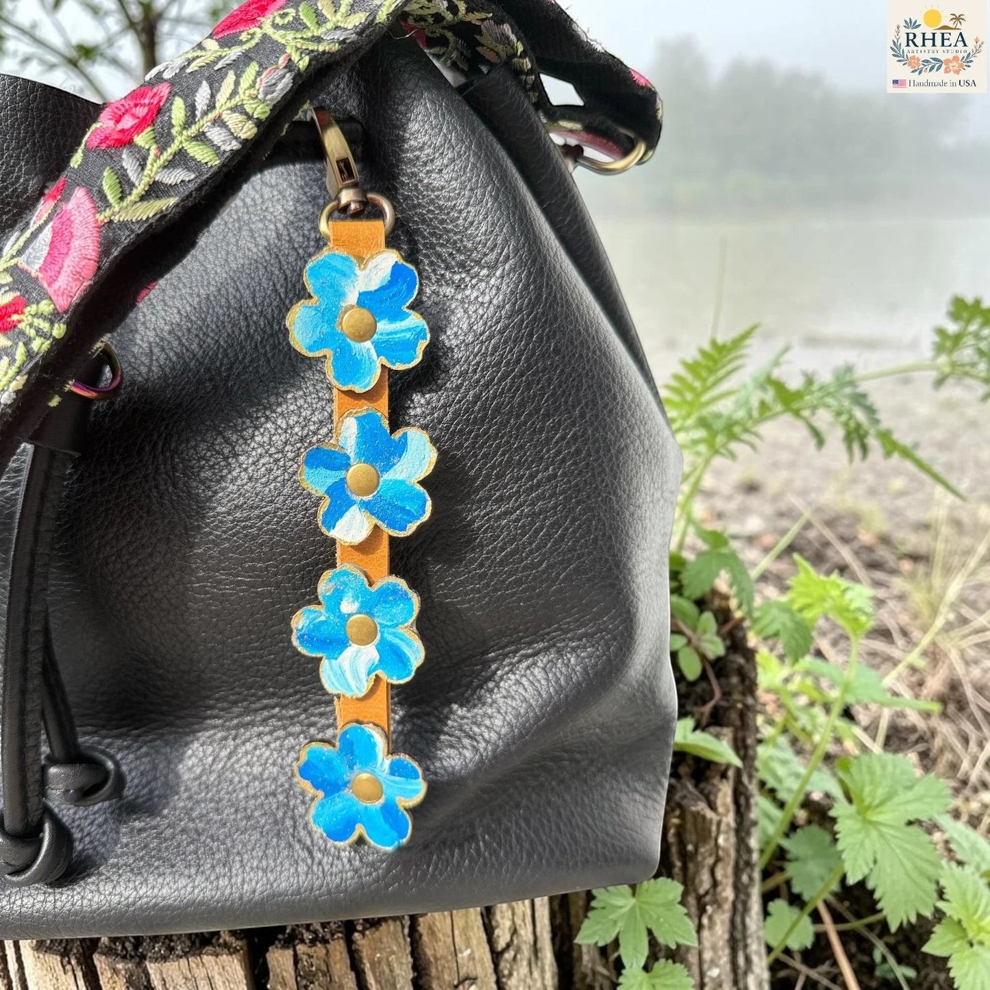 Rhea Artistry Studio Hand-Painted Leather Flower Bag Charm in Blue Sky with bright blue and white painted leather flowers, brass rivets, honey leather strap, and antique brass clasp.