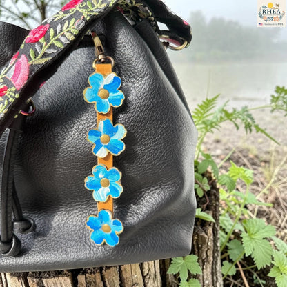 Rhea Artistry Studio Hand-Painted Leather Flower Bag Charm in Blue Sky with bright blue and white painted leather flowers, brass rivets, honey leather strap, and antique brass clasp.