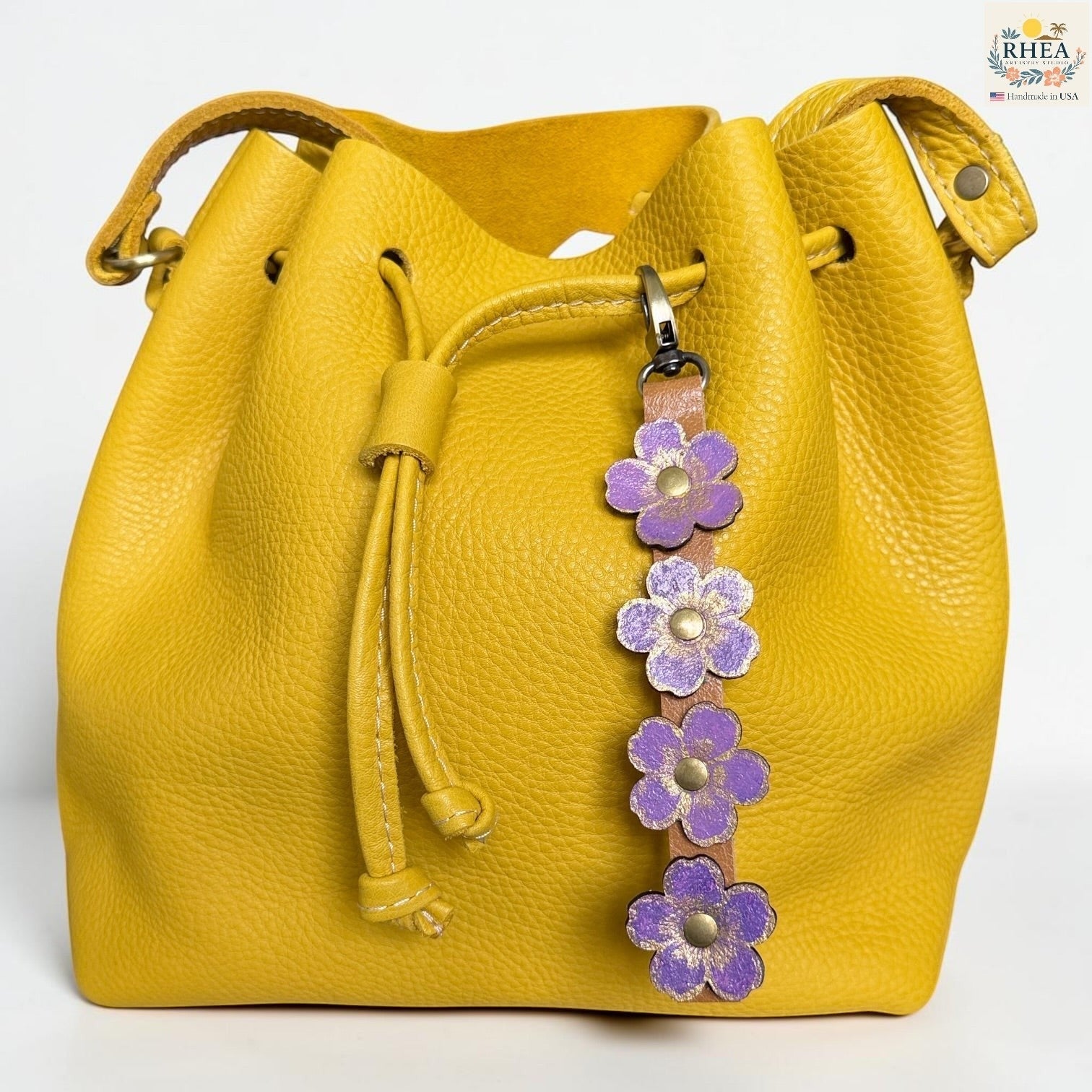 Rhea Artistry Studio Hand-Painted Leather Flower Bag Charm with lavender organic flower details, brass rivet centers, and antique brass hardware on a yellow bag