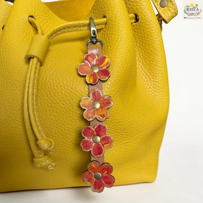 Rhea Artistry Studio Hand-Painted Leather Flower Bag Charm featuring Marigold Glow organic leather flowers with orange, coral, yellow, and brass details.