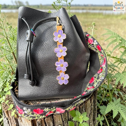 Rhea Artistry Studio Hand-Painted Leather Flower Bag Charm in Periwinkle Meadow with periwinkle blue and soft lavender painted leather flowers, brass rivets, honey leather strap, and antique brass clasp.