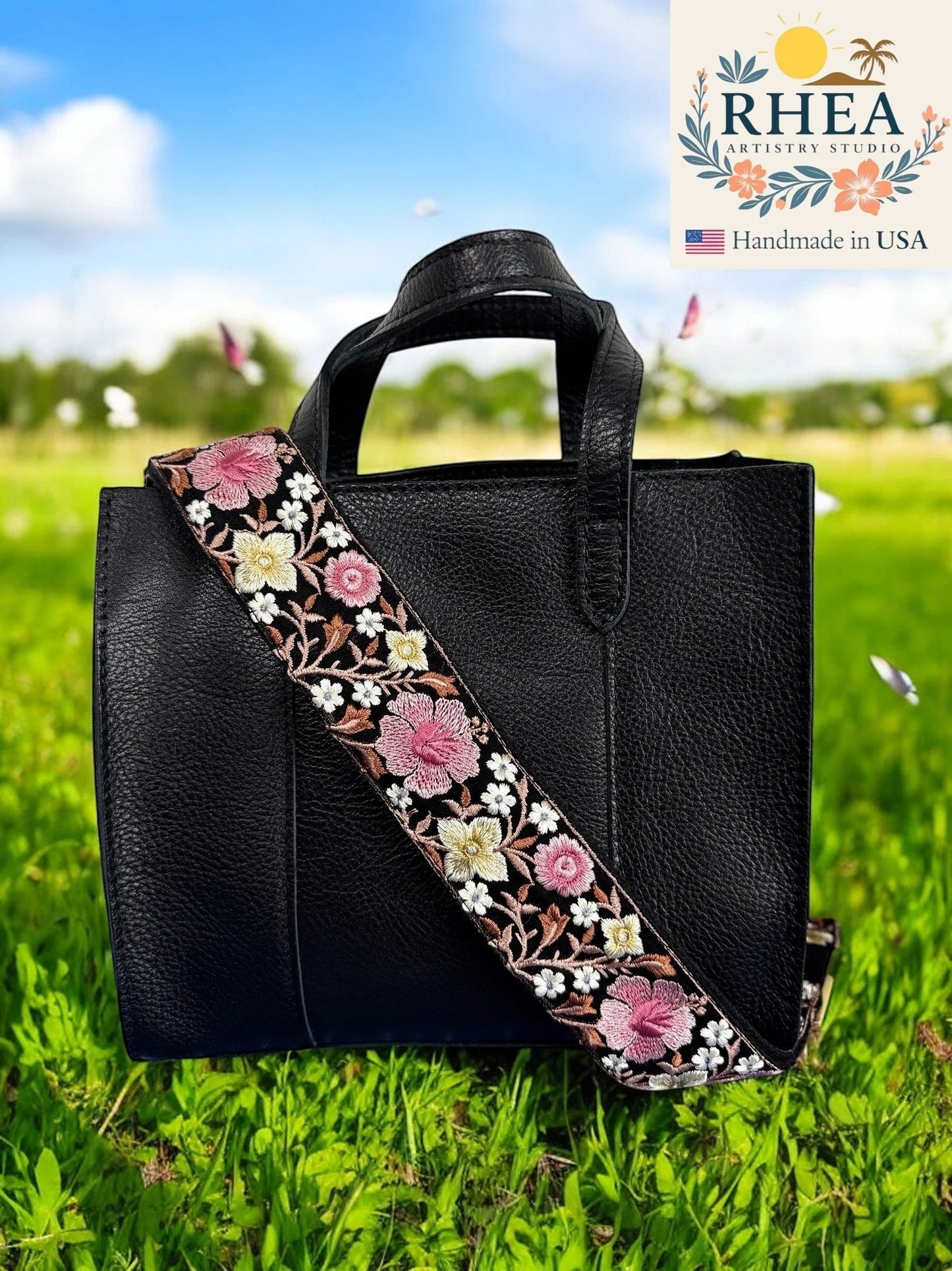 Rhea Artistry Studio 2-inch wide crossbody strap with pink and white hibiscus embroidery on black webbing, adjustable length, brushed brass hardware.