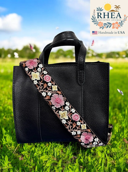 Rhea Artistry Studio 2-inch wide crossbody strap with pink and white hibiscus embroidery on black webbing, adjustable length, brushed brass hardware.