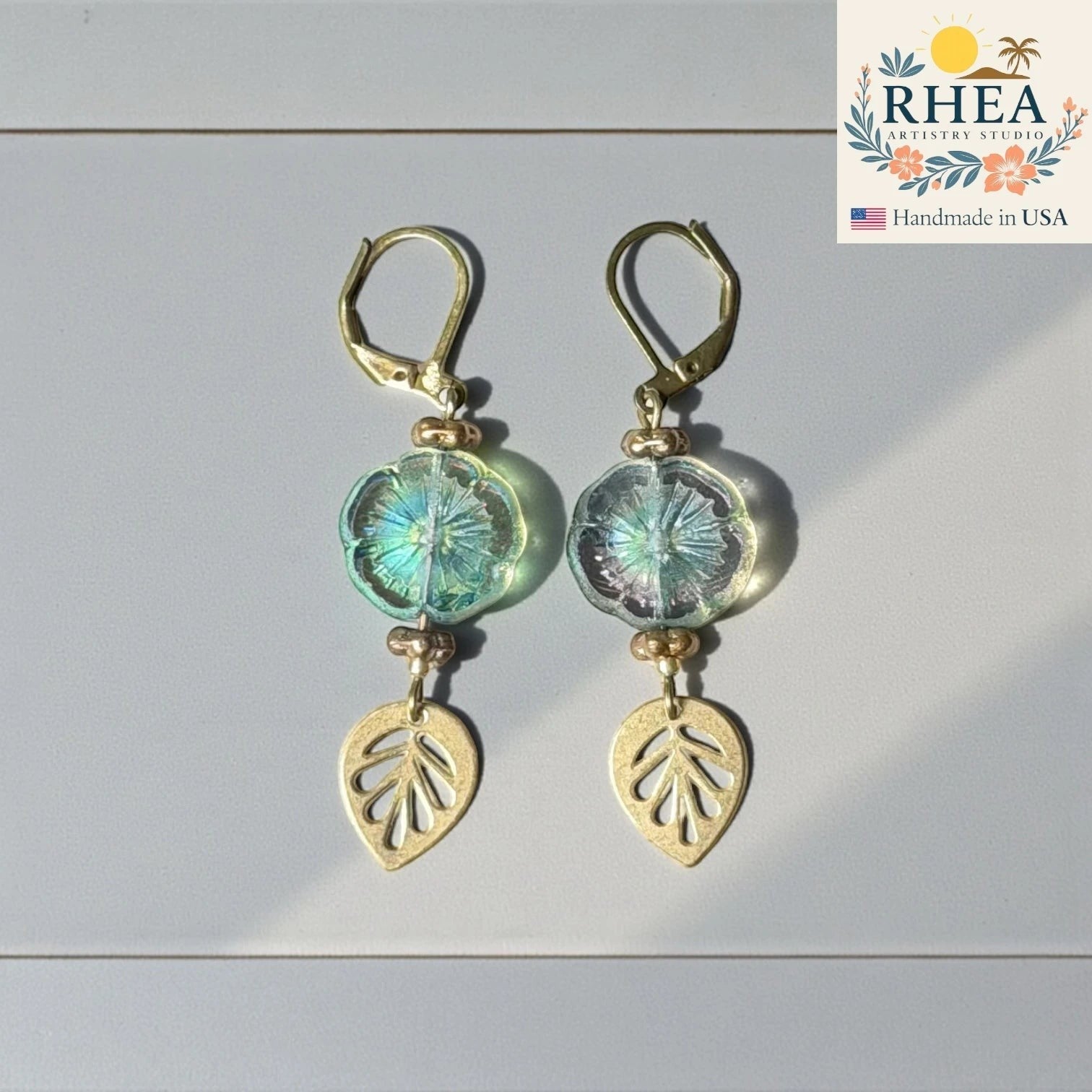 Rhea Artistry Studio handmade hibiscus glass drop earrings with gold leaf charms in iridescent blue-green.
