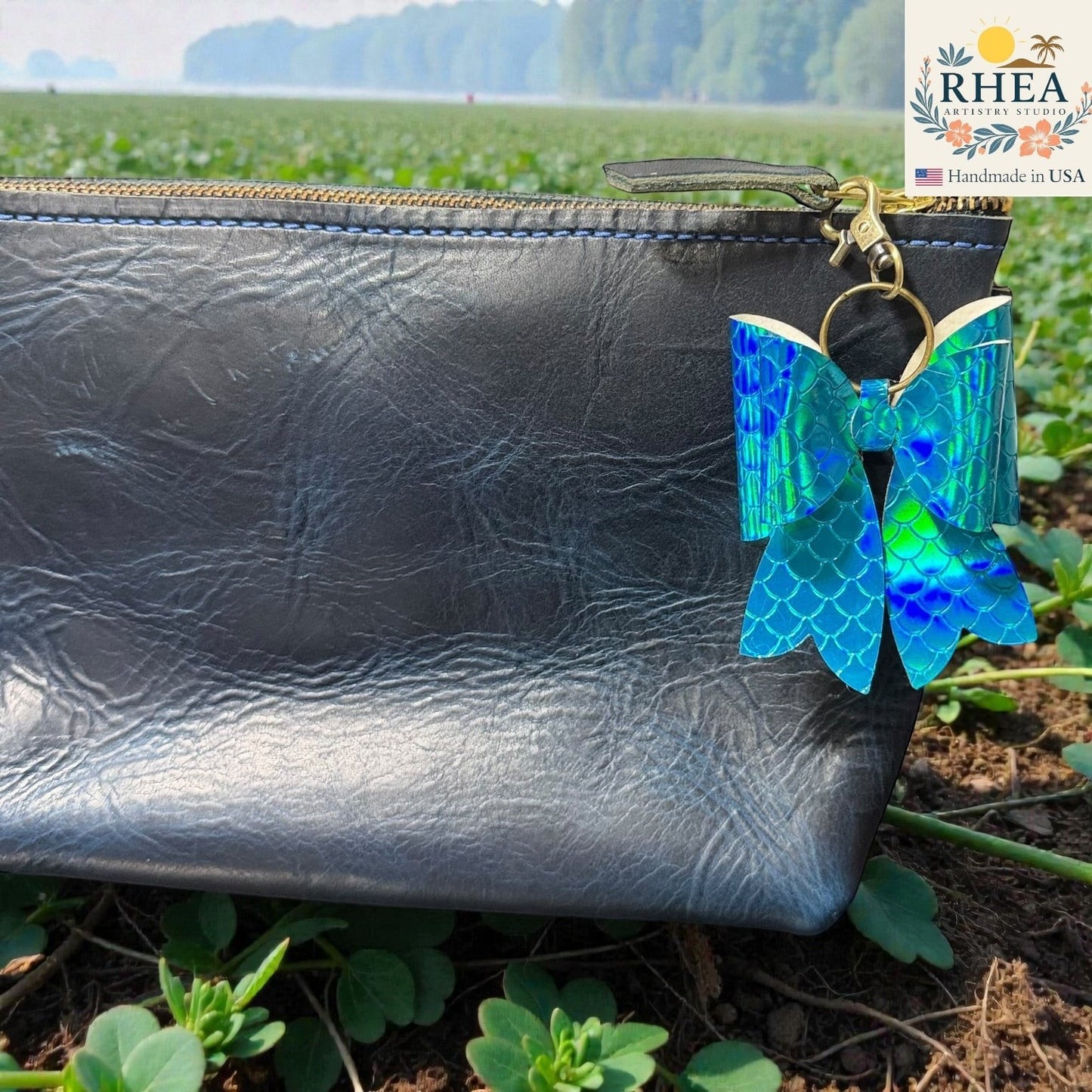 Rhea Artistry Studio holographic mermaid scale bow bag charm with brass hardware, attached to a dark grey clutch bag.