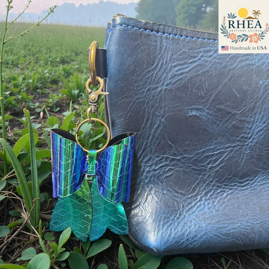 Rhea Artistry Studio holographic blue and green bow bag charm with brass hardware, attached to a dark blue leather bag.