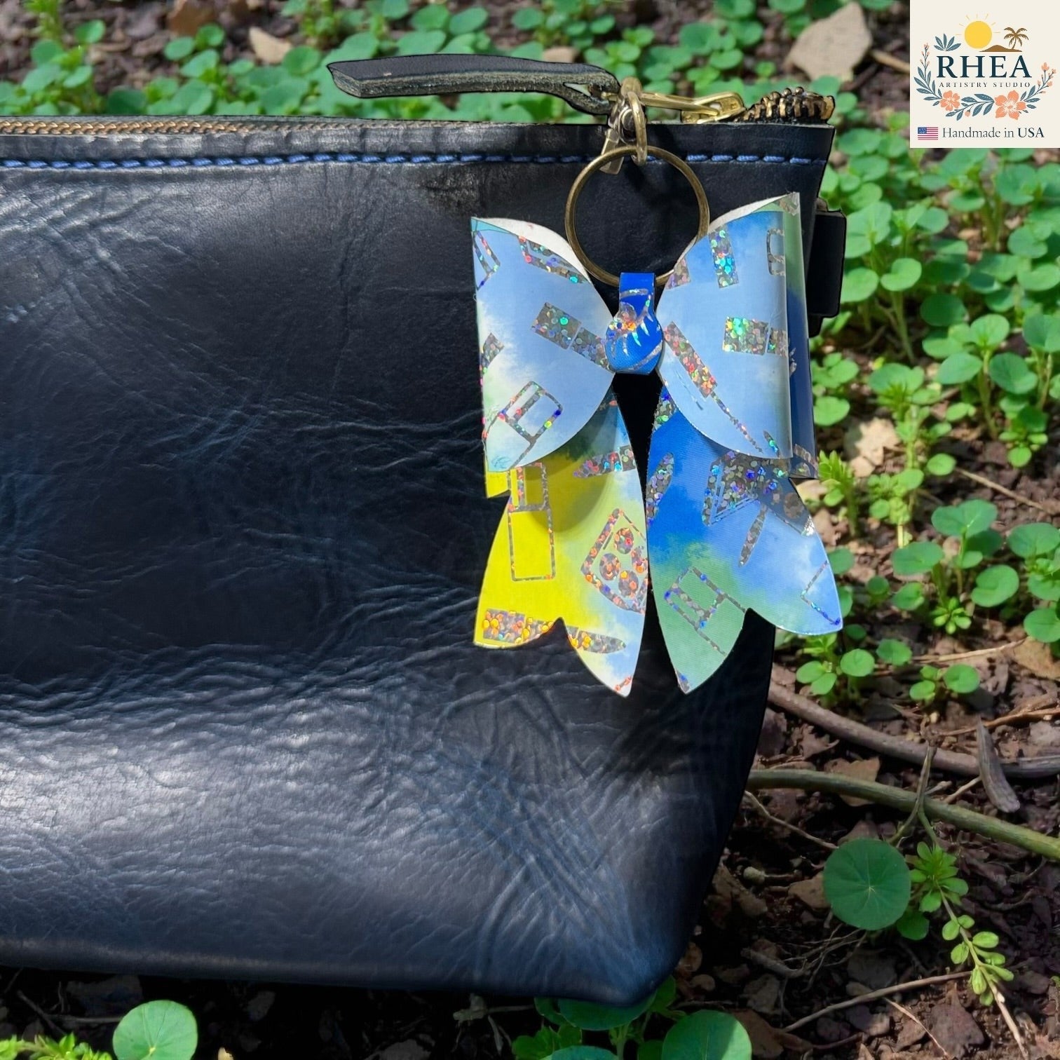 Rhea Artistry Studio holographic bow bag charm with blue, yellow, and green abstract print and glitter accents, attached to a black leather bag.