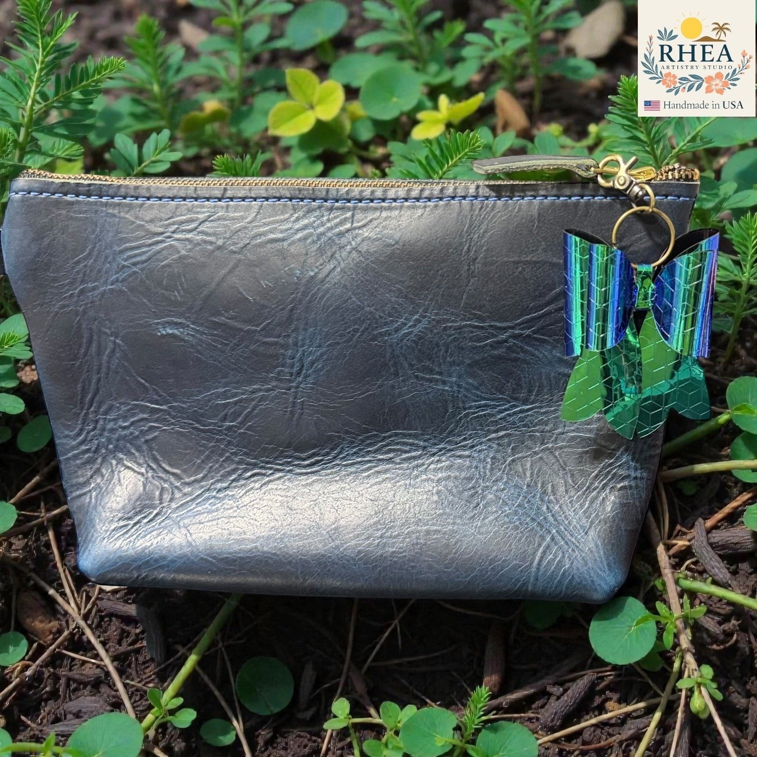 Rhea Artistry Studio holographic bow bag charm with green and blue iridescent finish, attached to a grey faux leather pouch.