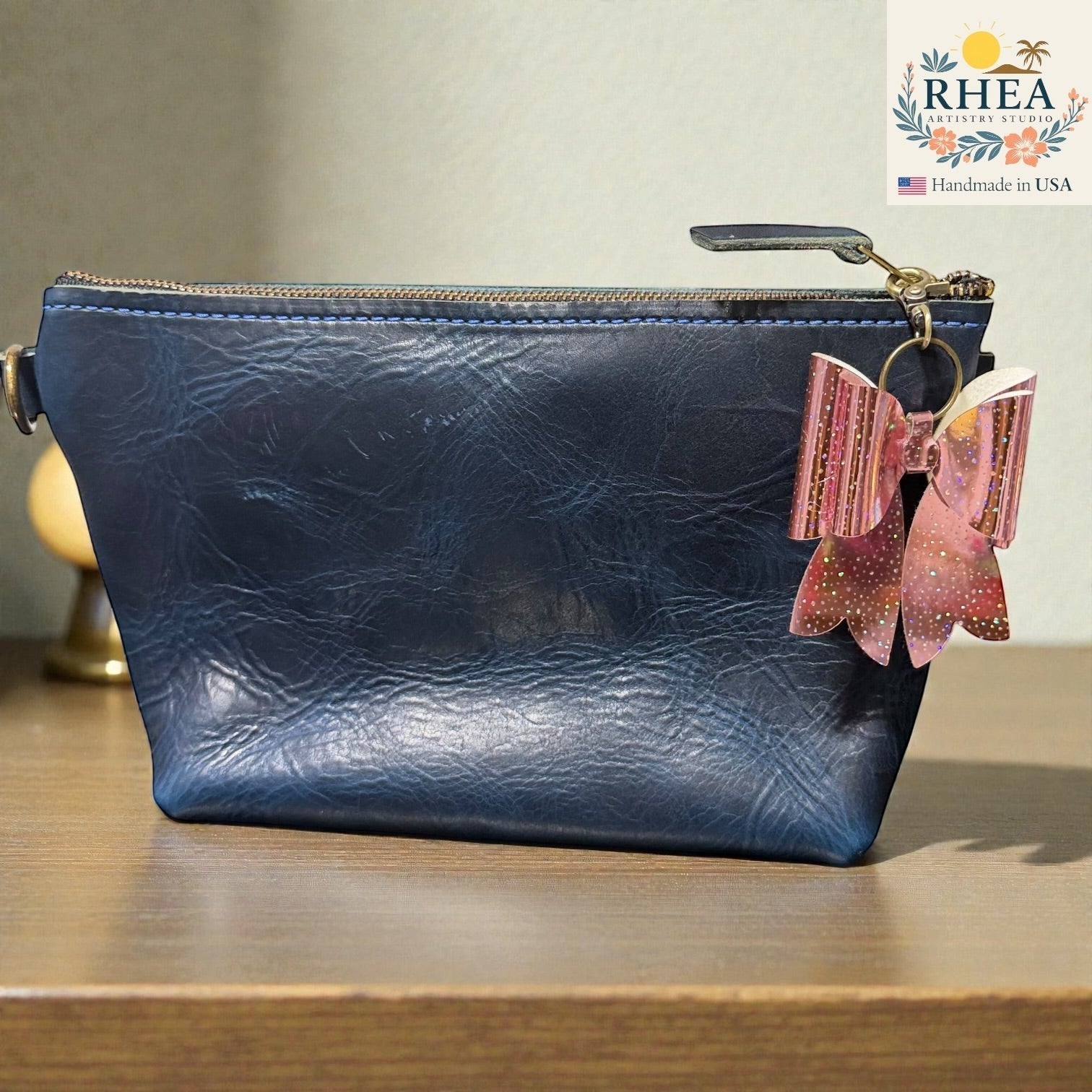 Rhea Artistry Studio holographic pink glitter bow bag charm attached to a navy blue leather pouch.