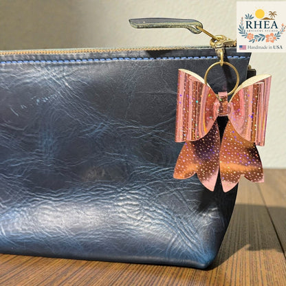 Rhea Artistry Studio holographic rose gold bow bag charm with glitter details and brass hardware attached to a navy blue leather pouch.