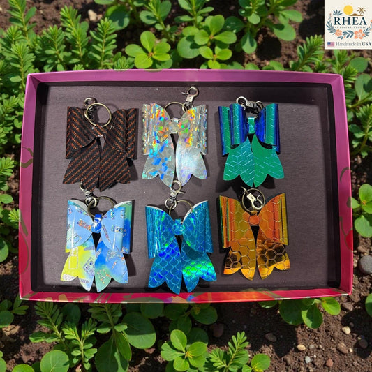 Rhea Artistry Studio collection of 6 holographic bow bag charms in various colors including iridescent white, blue, green, and orange, with brass hardware.