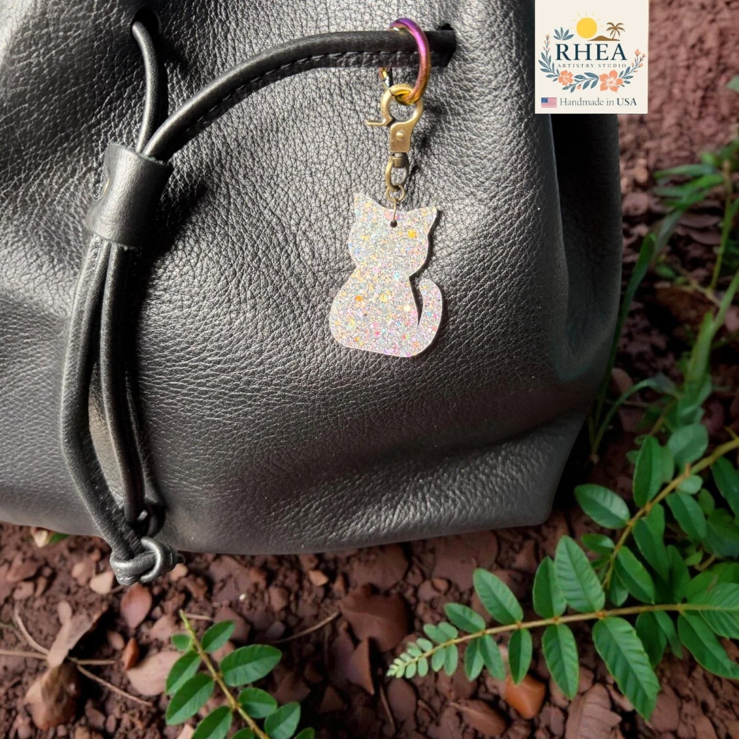 Rhea Artistry Studio holographic glitter cat silhouette acrylic charm with antique brass clasp on a black leather bag.