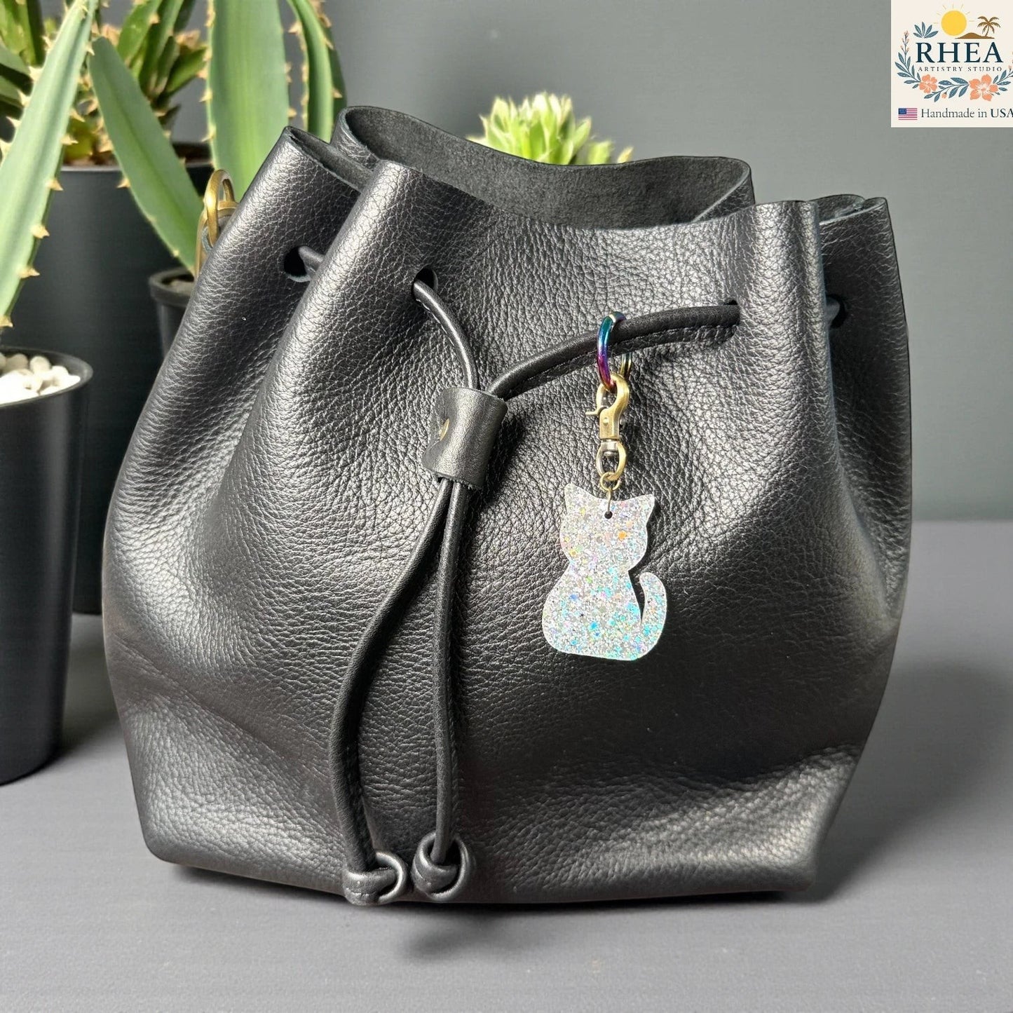Rhea Artistry Studio holographic glitter cat silhouette acrylic charm with antique brass clasp on a black leather drawstring bag.
