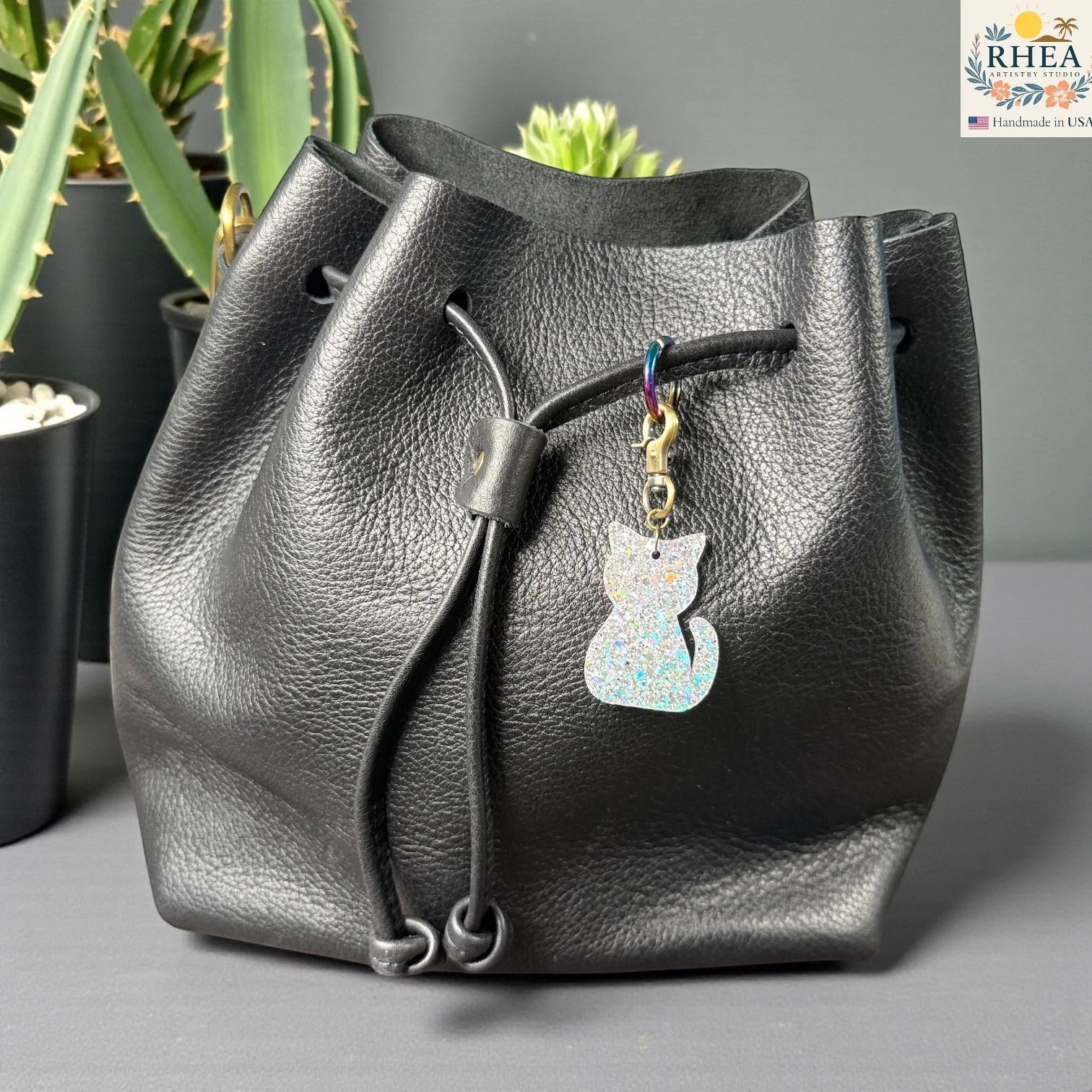 Rhea Artistry Studio holographic glitter cat silhouette acrylic charm with antique brass clasp on a black leather drawstring bag.