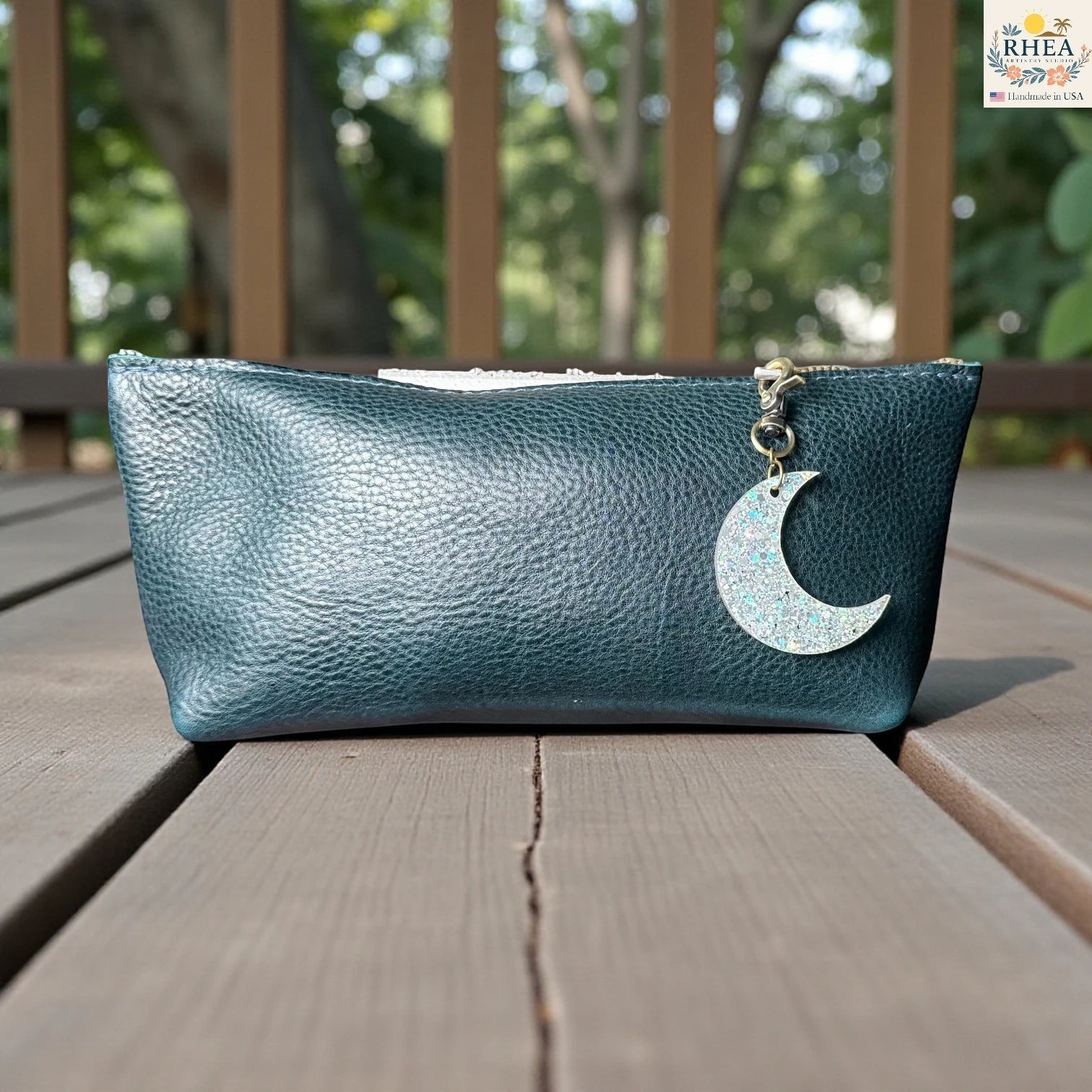 Rhea Artistry Studio holographic glitter moon charm on a teal leather pouch. The charm is crescent-shaped and hangs from a brass clasp.