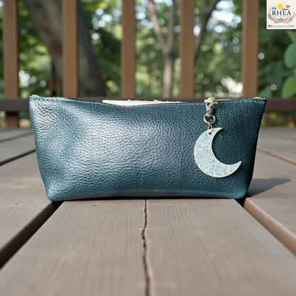 Rhea Artistry Studio holographic glitter moon charm on a teal leather pouch. The charm is crescent-shaped and hangs from a brass clasp.