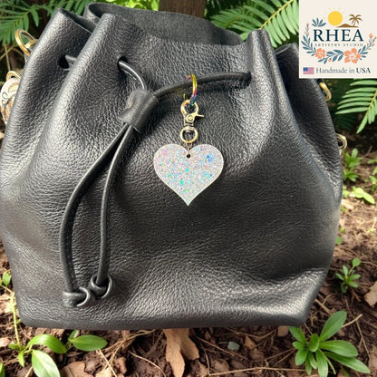 Rhea Artistry Studio holographic glitter heart charm with antique brass clasp on a black leather drawstring bag.