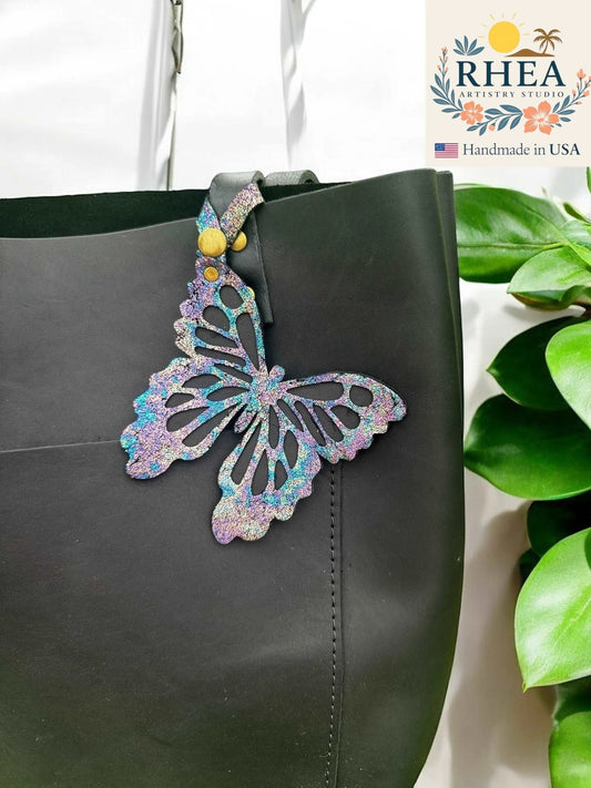 Rhea Artistry Studio iridescent leather butterfly charm with metallic finish attached to a black tote bag.